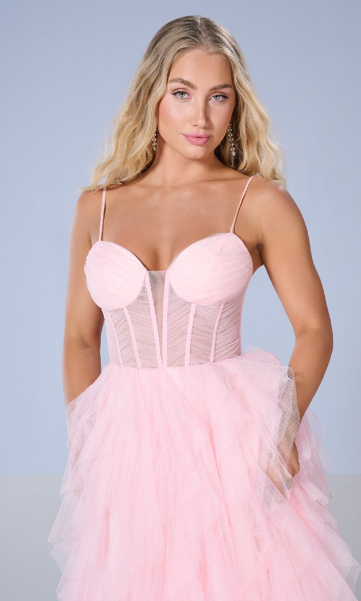 A woman with long blonde hair models the Tiffany 16161 Long Prom Dress, featuring a light pink corset bodice and layered tulle ball gown skirt, against a plain light blue background.