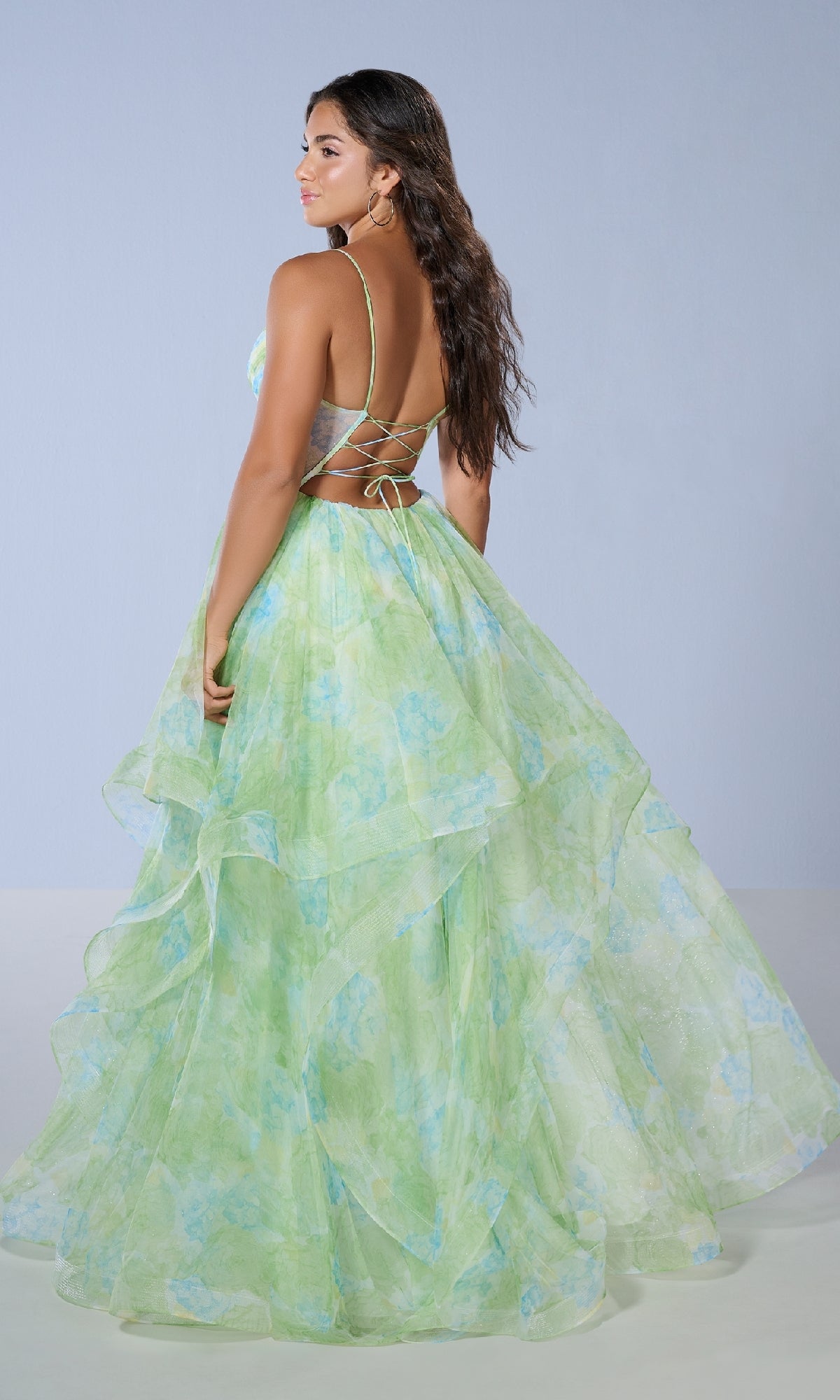 Woman wearing the Tiffany 16163 long prom dress featuring a green and blue floral print, open back, and crisscross straps, standing against a plain light background.