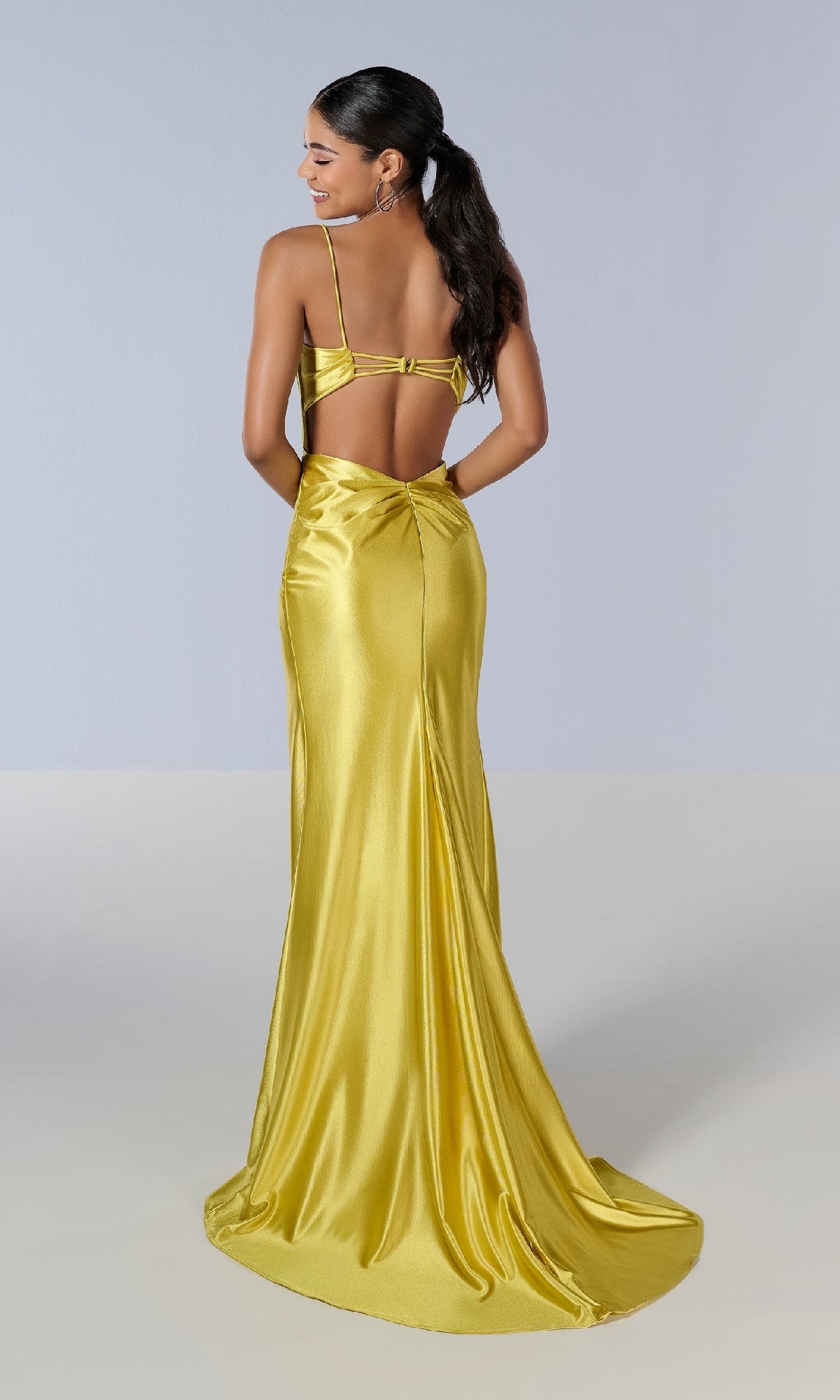 A woman stands against a plain light background wearing the Tiffany 16185 long prom dress, a shiny yellow satin gown featuring a floor-length train, open back, thin straps, and high slit.