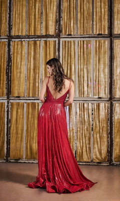 A woman wearing the Long Prom Dress: Tiffany 16189, a metallic pleated gown, stands with her back to the camera, facing gold curtains and a window framed by a rustic wooden grid.