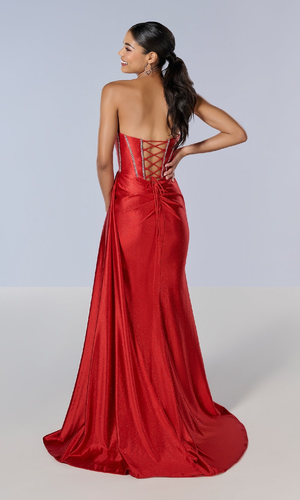 Woman wearing the Tiffany 16197 long prom dress, featuring a strapless glitter design, lace-up back, and floor-length skirt, stands with one hand on her hip, facing away from the camera.