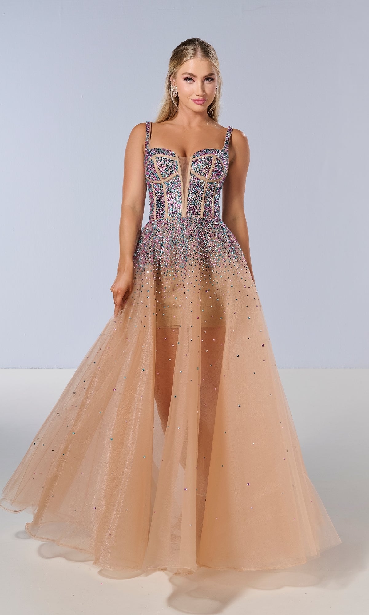 A woman models the Tiffany 16206 long prom dress—a sleeveless, sheer peach tulle gown with a beaded, colorful corset bodice, crystal details, and a deep neckline—posing against a plain light background.