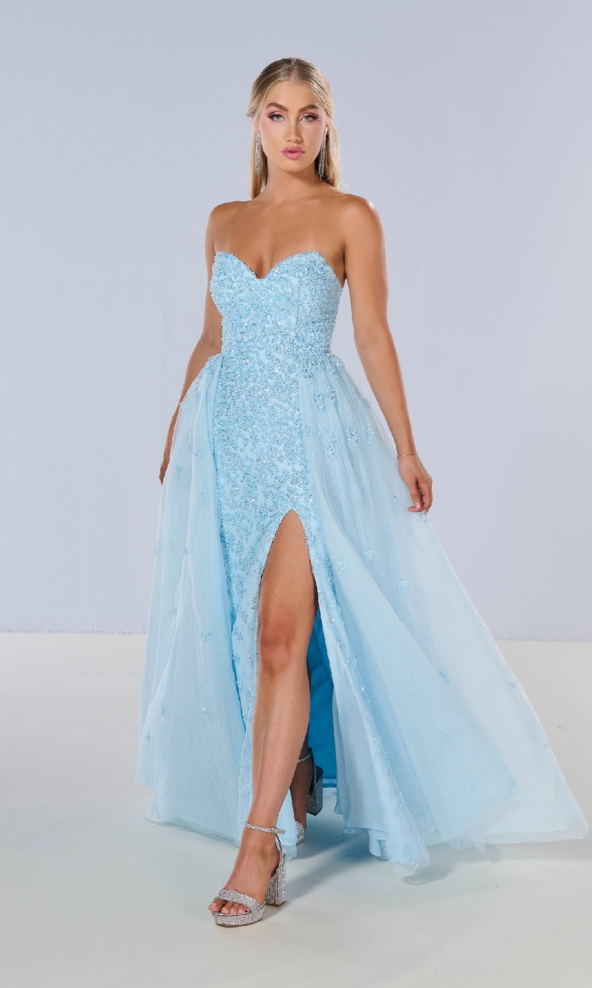 A woman models the Tiffany 16209 long prom dress—light blue, strapless with a sweetheart neckline, thigh-high slit, floral details—and silver high-heeled sandals, set against a plain light background.