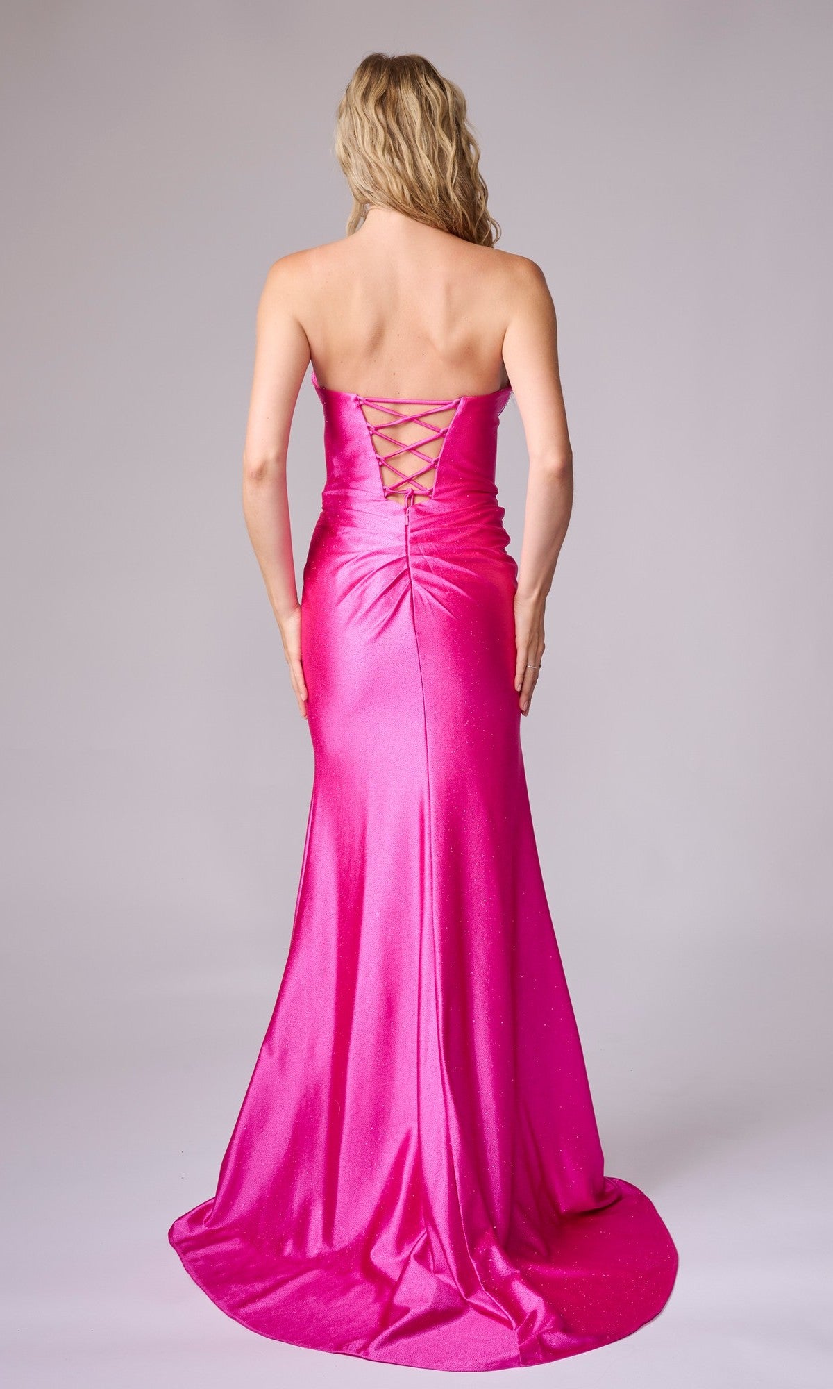 A woman models the Nina Canacci 1620, a strapless long formal dress in bright pink with a lace-up back, standing with her back to the camera against a plain backdrop.