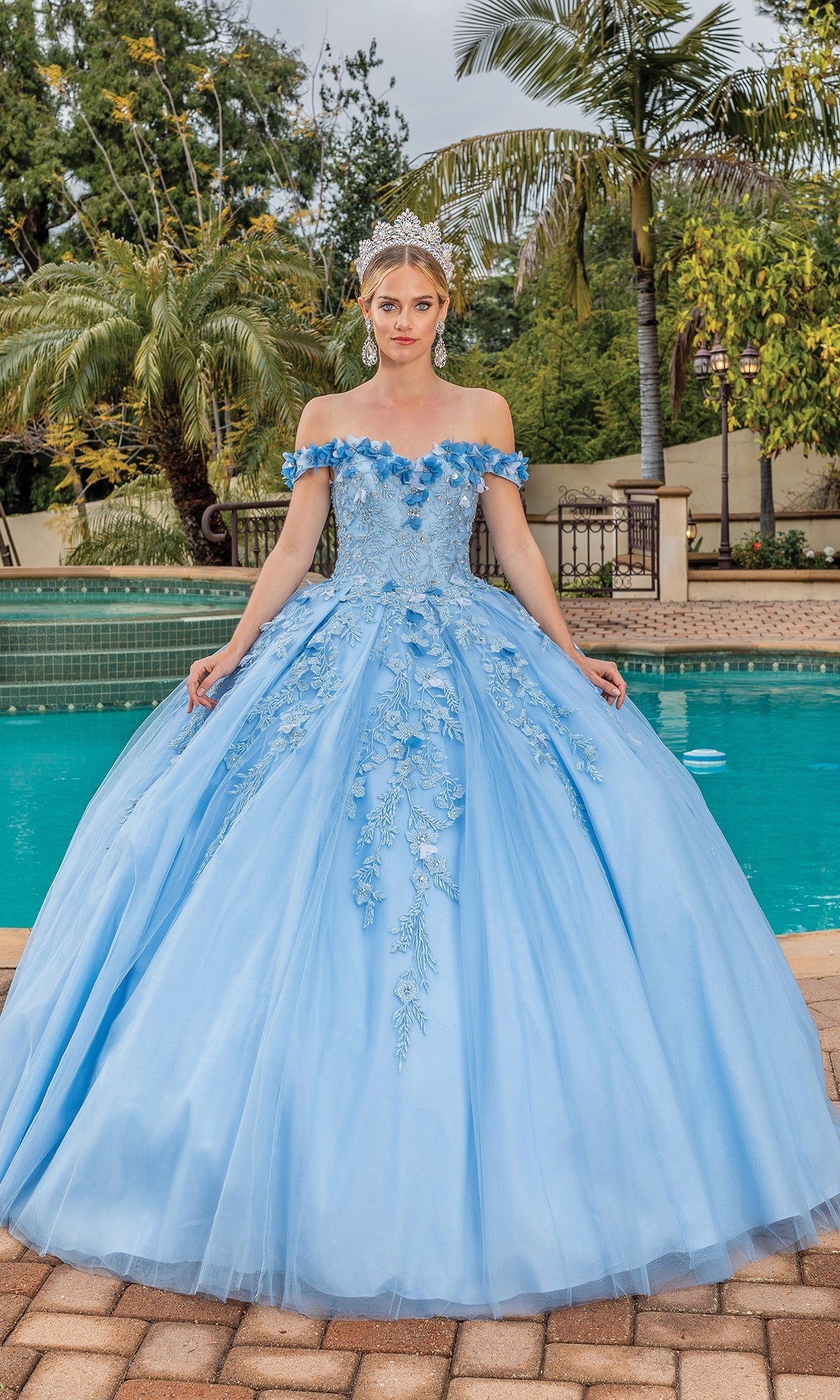 A woman stands by a pool surrounded by palm trees, wearing the Quinceanera Dress 1620 By Dancing Queen—a light blue, off-the-shoulder gown with floral details and a sweetheart neckline.