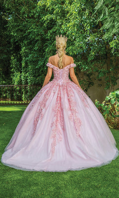 A woman wearing the Quinceanera Dress 1620 By Dancing Queen, a light pink off-the-shoulder gown with lace details and a crown, stands on grass facing away, with trees in the background.