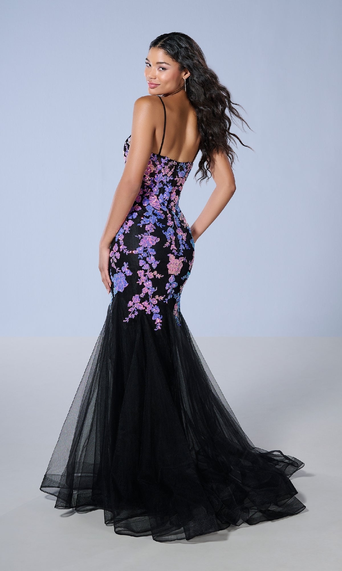 Woman wearing Tiffany 16240 long prom dress—a black fitted mermaid gown with pink and purple floral sequin appliqué, sheer tulle bottom, and thin straps—poses with her back turned, glancing over her shoulder.