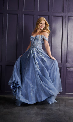 A young woman wears the Tiffany 16243 Long Prom Dress with glitter tulle, standing before a dark purple paneled wall. She smiles as she holds the sides of her sparkling, off-the-shoulder gown.
