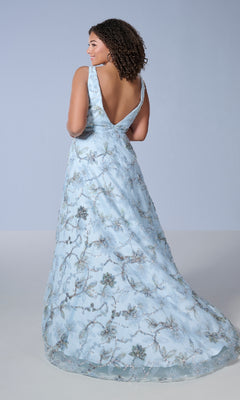 A woman showcases the Long Plus-Size Prom Dress Tiffany 16249, featuring light blue fabric with silver floral embroidery, a deep V-back, and elegant embellished details.