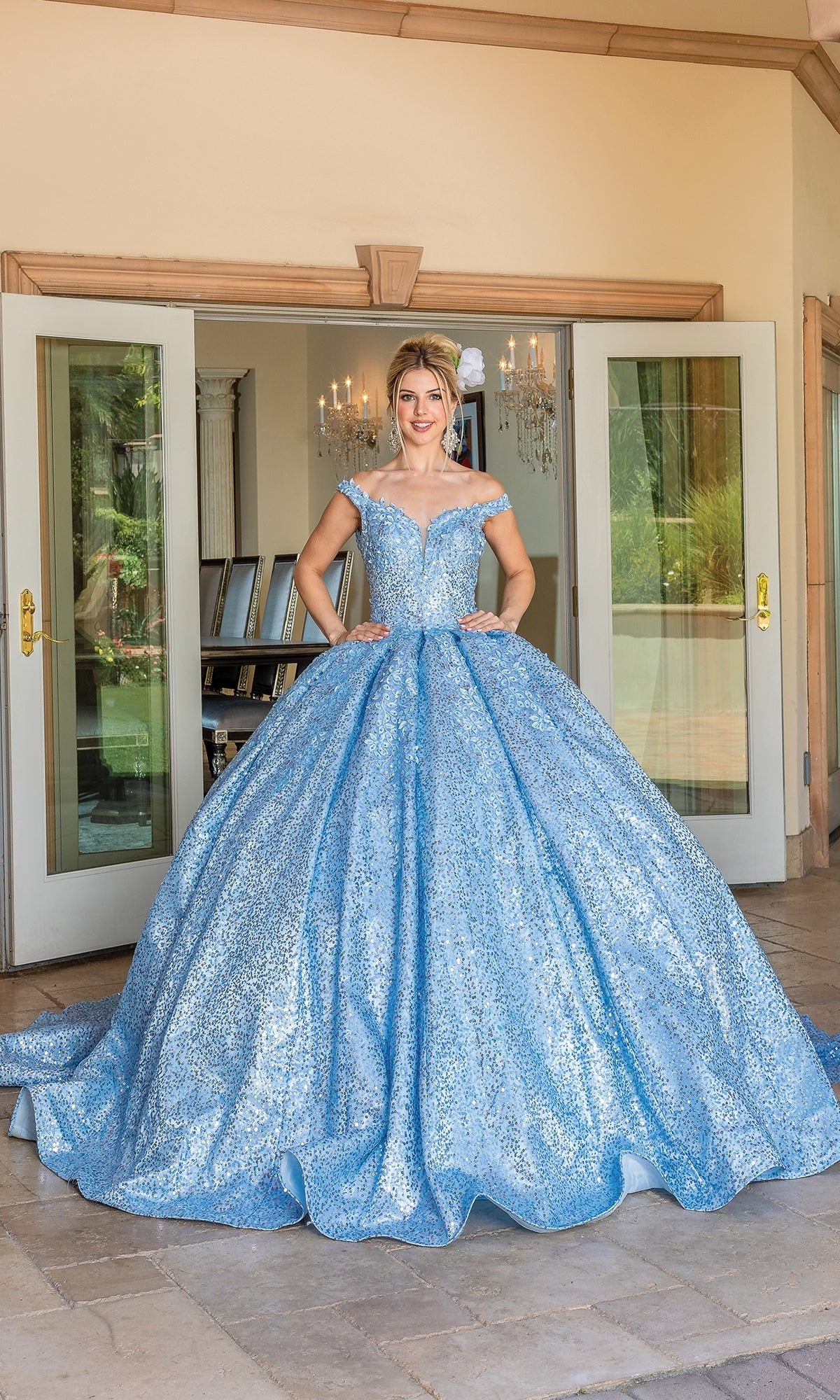 A person stands before open double doors wearing the Quinceanera Dress 1624 By Dancing Queen, featuring an off-the-shoulder design, sparkling full skirt, and elegant sweetheart neckline.