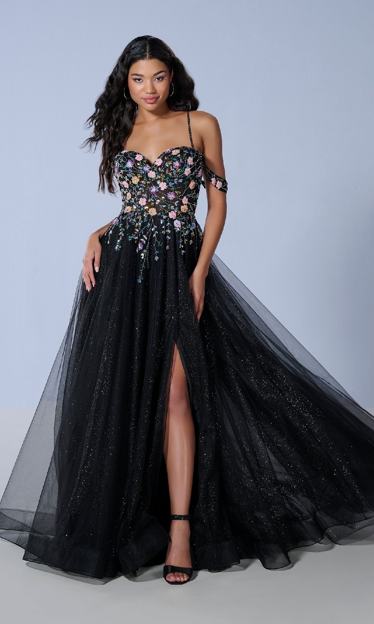 A woman stands against a plain light background wearing the Tiffany 16251 long prom dress—a black, floor-length A-line gown with floral beaded applique, off-the-shoulder straps, and a front leg slit.