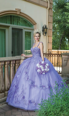 A woman in the Quinceanera Dress 1627 By Dancing Queen—purple with floral details and a crown—stands on a balcony holding a matching bouquet, with greenery and a building in the background.