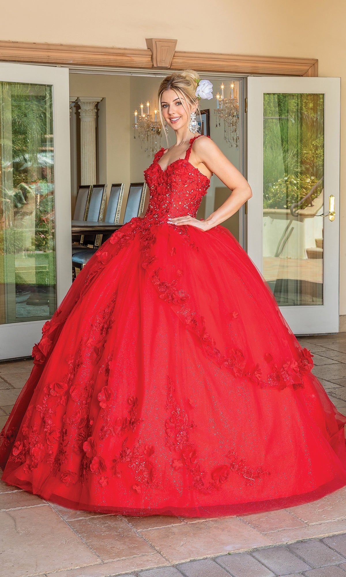 A woman poses outdoors in front of open glass doors, wearing the Quinceanera Dress 1627 By Dancing Queen—a bright red ball gown with a sweetheart neckline, full skirt, and floral details.
