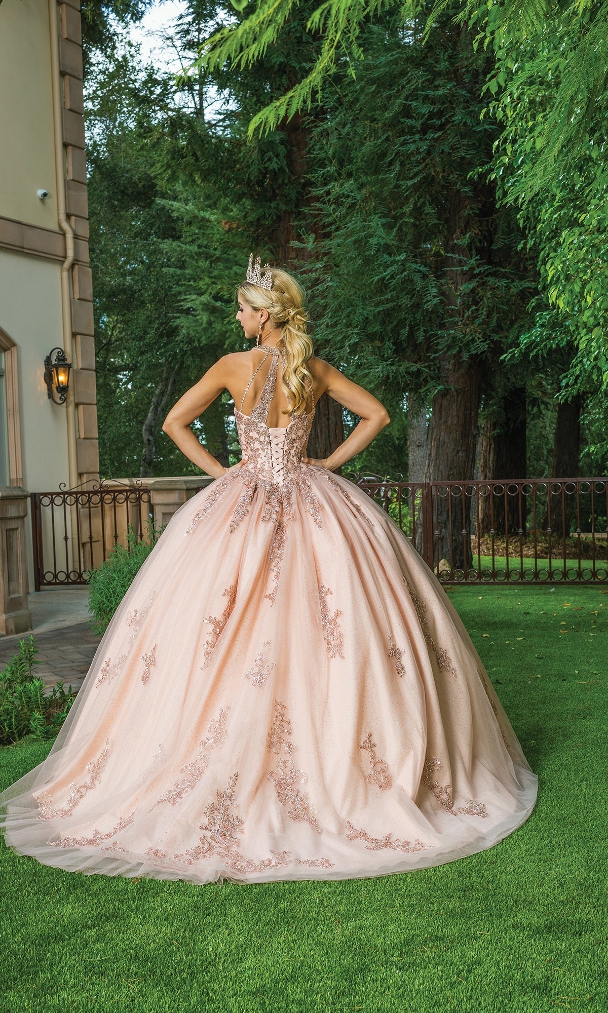 A woman in the Rose Gold Quinceanera Dress 1628, featuring floral embroidery, stands on grass with a crown, facing away near a building and tall trees.