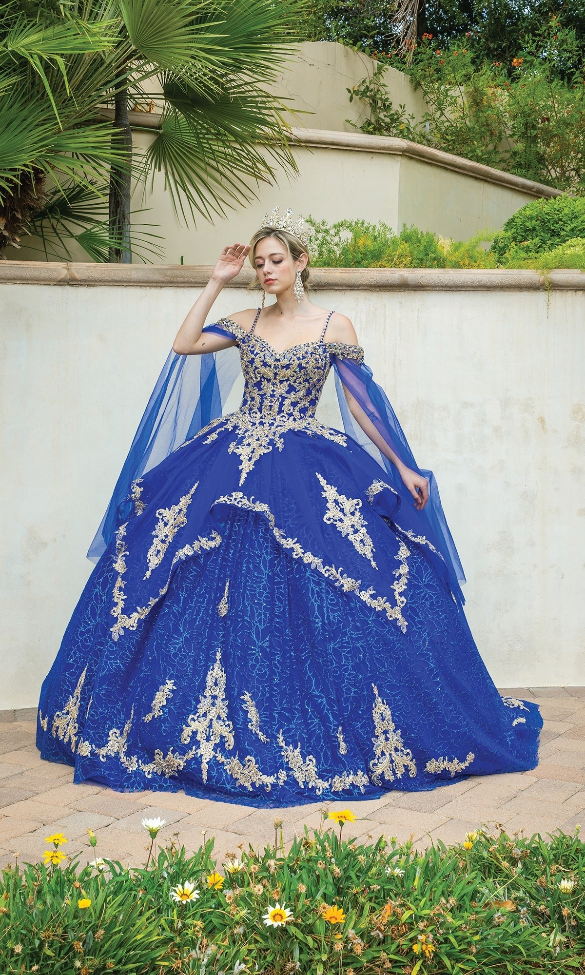 A person wearing the Quinceanera Dress 1638 By Dancing Queen, an ornate royal blue off-the-shoulder ball gown with gold detailing and a tiara, stands outdoors near a white wall and greenery.