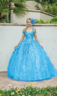 A woman stands outdoors in front of a white wall, wearing the Quinceanera Dress 1640 by Dancing Queen—an off-the-shoulder A-line ball gown with bright blue floral details and a matching floral headpiece.