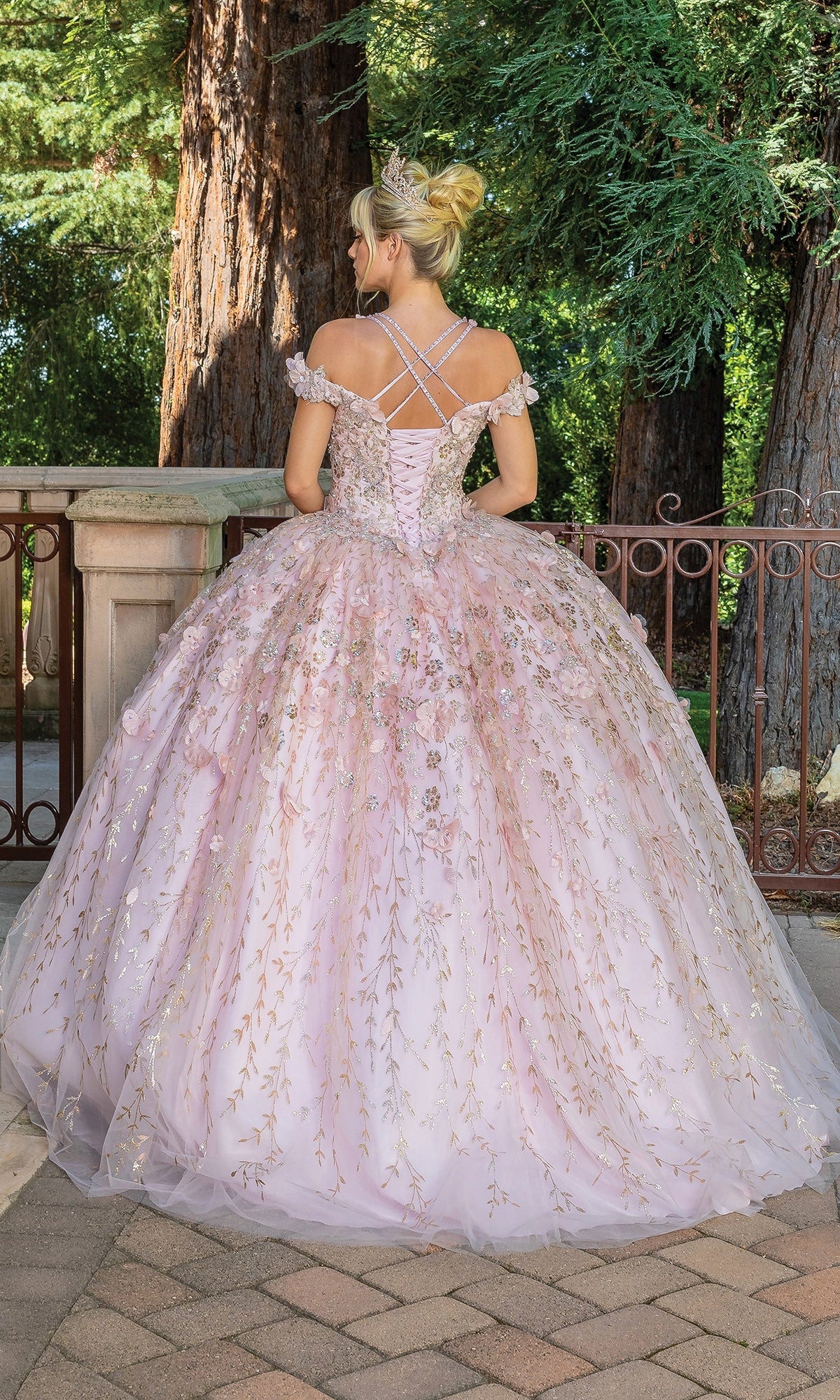 Someone wearing the Quinceanera Dress 1640 By Dancing Queen—a pink off-the-shoulder a-line ball gown with gold floral accents and a tiara—stands outdoors with their back to the camera.