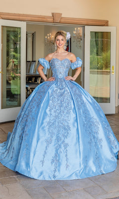 A young woman stands indoors wearing the Quinceanera Dress 1644 By Dancing Queen, a light blue strapless ball gown with intricate embroidery, a full skirt, and removable puff sleeves.