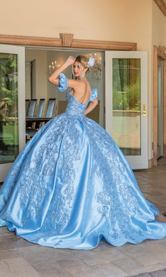 A woman stands outside double doors wearing Quinceanera Dress 1644 by Dancing Queen, a voluminous light blue A-line gown with floral embroidery and off-the-shoulder sleeves, facing slightly away from the camera.