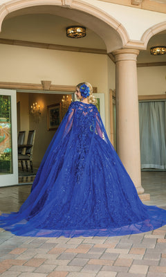 A woman stands outdoors before an ornate building, wearing Quinceanera Dress 1649 By Dancing Queen—a royal blue ball gown with a sweetheart neckline, long train, and matching headpiece.