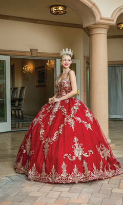 A woman in the Quinceanera Dress 1651 by Dancing Queen, a red A-line ball gown with gold embroidery and a sweetheart neckline, stands in an elegant foyer near French doors, wearing a jeweled crown.