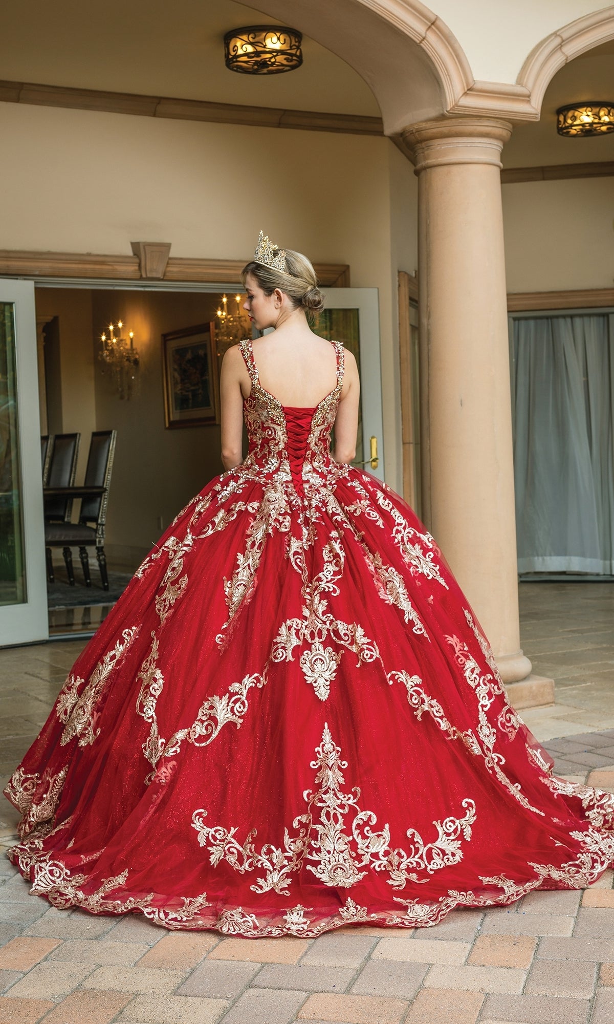 A person wearing the Quinceanera Dress 1651 by Dancing Queen, featuring red and gold lace with a sweetheart neckline and tiara, stands facing away in front of an arched entryway.