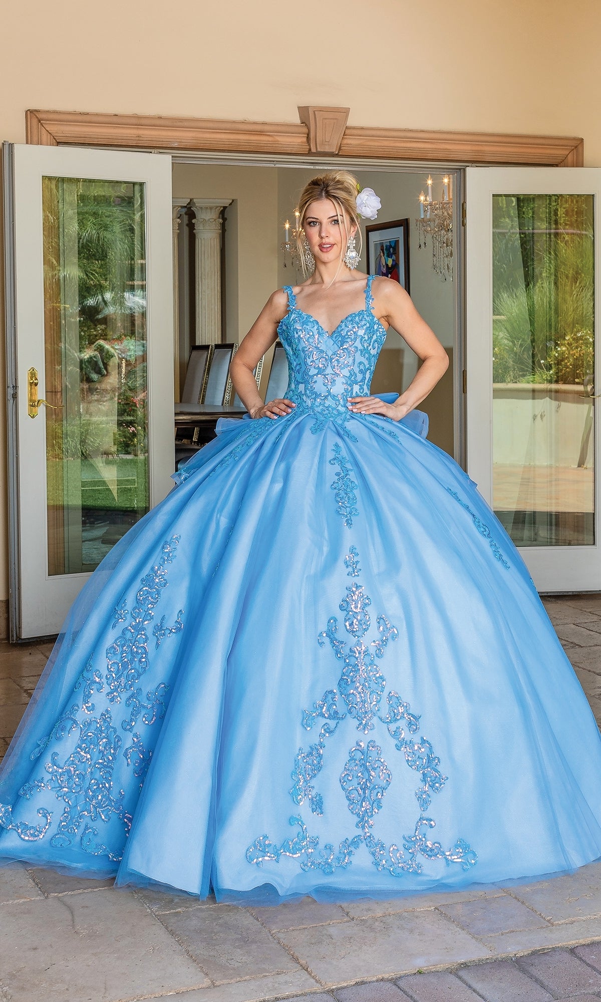 A woman stands in a doorway wearing the Quinceanera Dress 1652 By Dancing Queen, a light blue A-line ball gown with silver embroidery and a tiara, posing with hands on her waist.