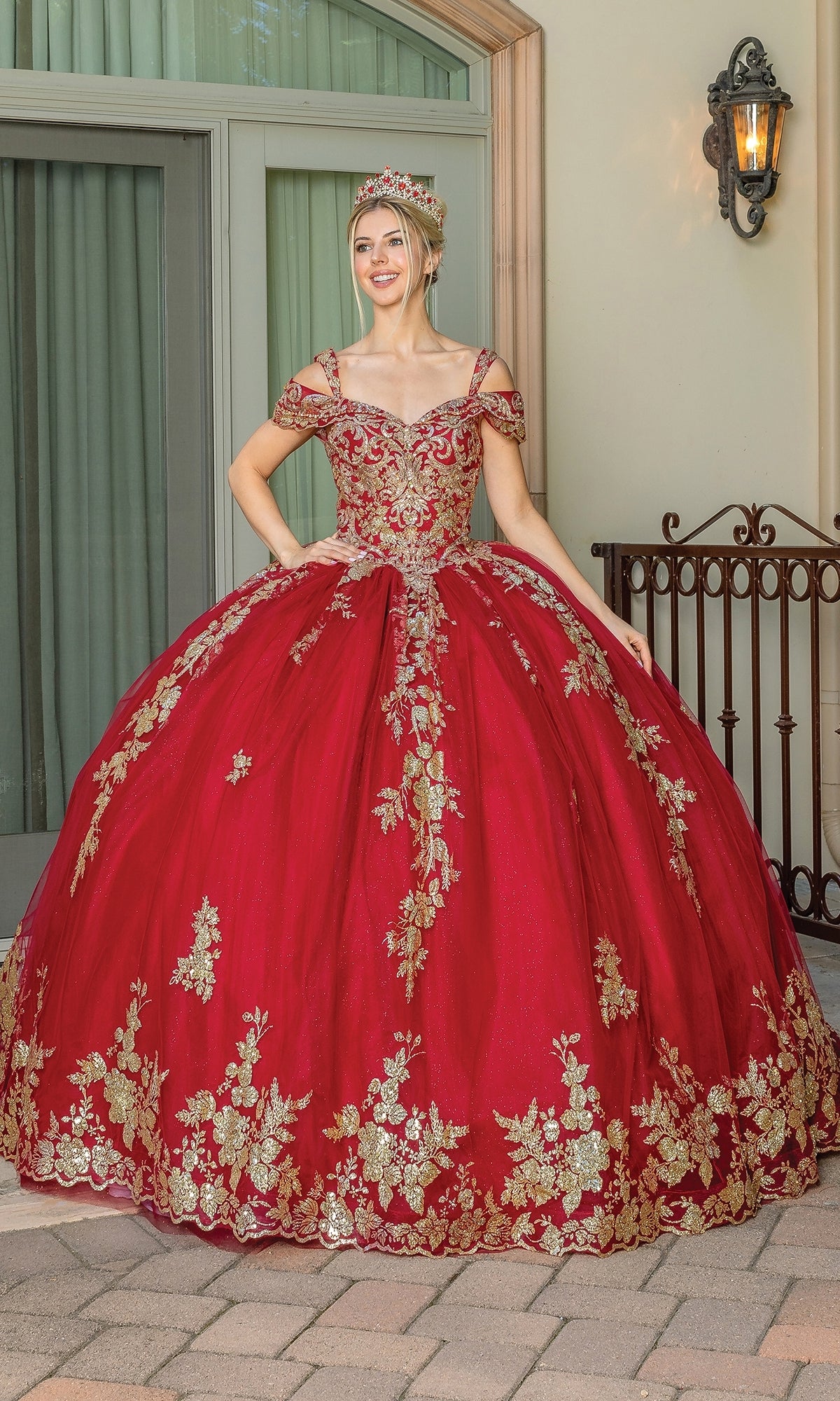 A woman poses outdoors in the Burgundy Red Quinceanera Dress 1657, featuring gold floral embroidery and a matching crown, smiling brightly at the camera.