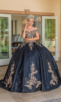 A woman wearing the Quinceanera Dress 1658 By Dancing Queen, an off-the-shoulder ball gown with gold embroidery and a jeweled crown, stands in front of open glass doors.