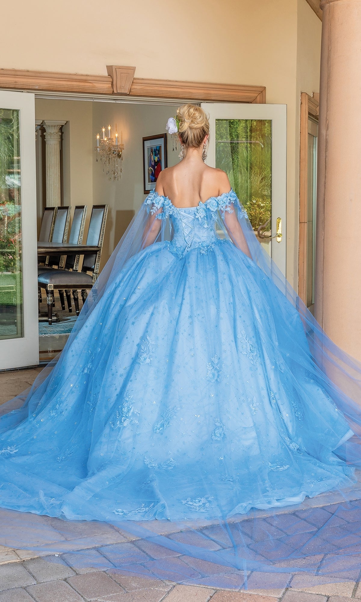A woman with a flower in her hair wears the Quinceanera Dress 1660 By Dancing Queen, an off-the-shoulder gown with a long train, standing before open glass doors.