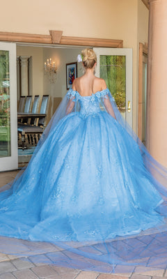 A woman with a flower in her hair wears the Quinceanera Dress 1660 By Dancing Queen, an off-the-shoulder gown with a long train, standing before open glass doors.