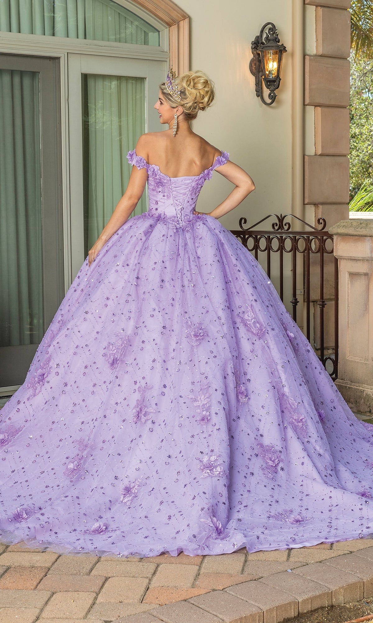 A woman stands outdoors facing away from the camera, wearing the Quinceanera Dress 1660 by Dancing Queen with floral embroidery, removable cape sleeves, and a tiara in front of a building.