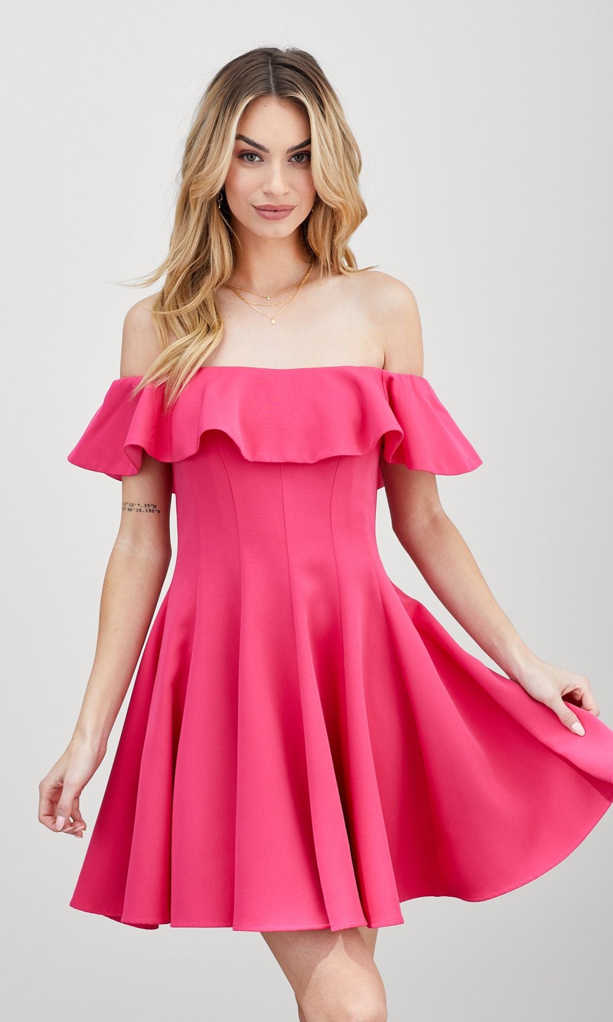 Model wearing the Off-the-Shoulder Short Party Dress in bright pink with ruffles, holding the skirt’s sides while standing against a plain light background.