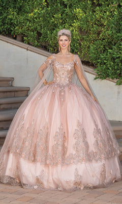 A woman in the Dancing Queen 1676 Quince Dress with Cape Sleeves, featuring ornate blush pink embroidery and a tiara, stands on a paved path before green foliage and stairs.