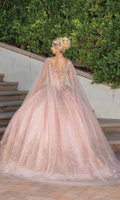 A woman in the Dancing Queen 1676 Quince Dress with Cape Sleeves, a blush pink embellished ball gown, stands outdoors on paved steps before greenery, facing away and evoking the elegance of a classic corset quinceañera look.