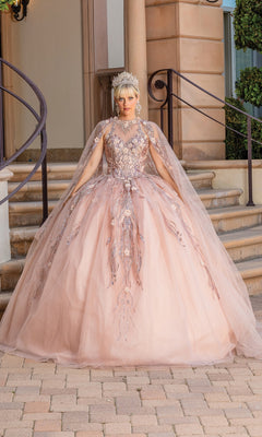 A person wears the Sleeveless Quinceanera Dress 1677, a pale pink ball gown with intricate floral embroidery and a removable sheer cape, paired with a jeweled crown, standing on stone steps in front of a building.