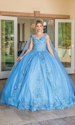 A woman stands before glass doors in the Quinceanera Dress 1678 By Dancing Queen, a blue ball gown with floral lace, removable draped sleeves, and a full skirt, styled with an updo and hair accessory.