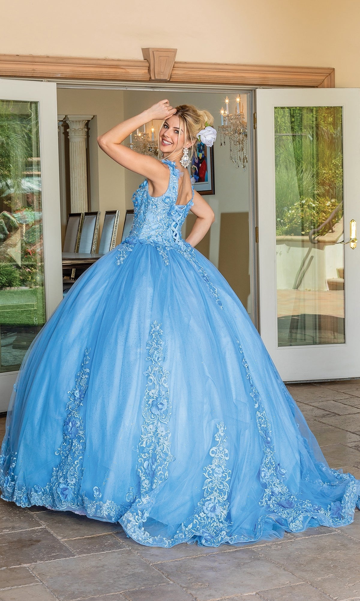 A woman in the Quinceanera Dress 1678 by Dancing Queen, featuring blue floral details and removable draped sleeves, stands before open glass doors, smiling over her shoulder.