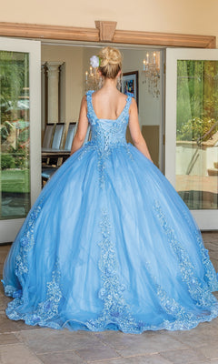 A woman with blonde hair in an updo wears the Quinceanera Dress 1678 By Dancing Queen, a floor-length light blue A-line ball gown with lace details and removable draped sleeves, standing facing glass doors from behind.