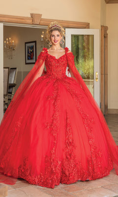 A woman stands indoors wearing the Quinceanera Dress 1678 by Dancing Queen, a bright red ball gown with floral details, matching cape, removable draped sleeves, plus a tiara and earrings.