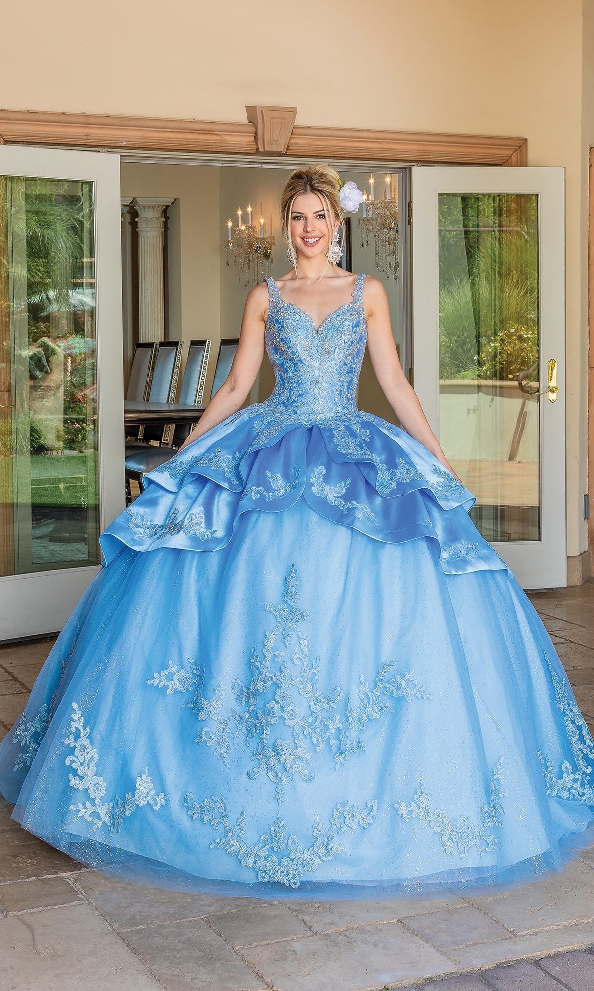 A young woman smiles in front of open glass doors, wearing the Quinceanera Dress 1682 By Dancing Queen—a light blue ball gown with layered tulle, a sweetheart neckline, and delicate floral embroidery.