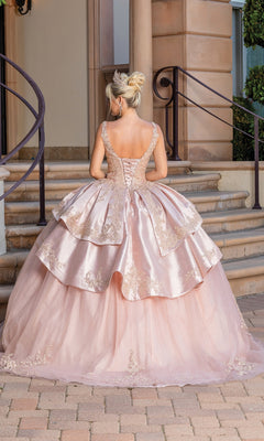 A woman with blonde hair in a bun wears Quinceanera Dress 1682 by Dancing Queen, a pink layered ball gown with lace details and sweetheart neckline, standing on a paved area before stone steps and a building.