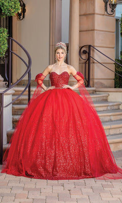 A woman stands on stone steps in the Quinceanera Dress 1683 By Dancing Queen, a sparkling red ball gown with off-the-shoulder sleeves and a sweetheart neckline, posing in front of a building entrance while wearing a tiara.