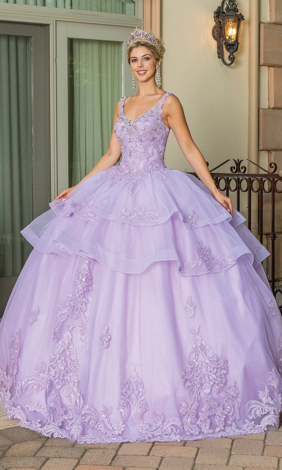A woman in the Quinceanera Dress 1686 By Dancing Queen, featuring lilac fabric and lace details, stands outside a building entrance wearing a jeweled tiara.