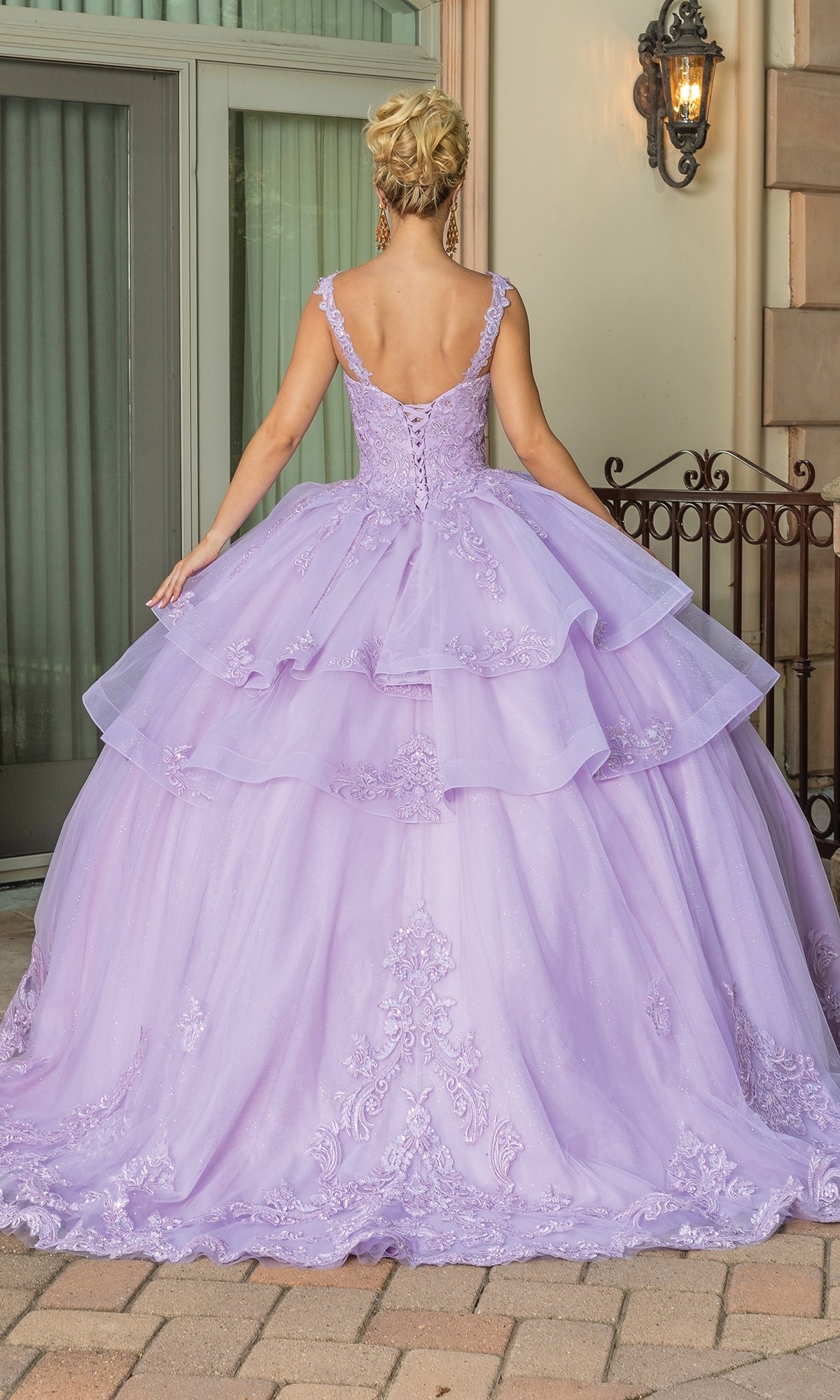 Woman wearing Quinceanera Dress 1686 By Dancing Queen, an elaborate lilac gown with layered tulle and embroidered details, standing outdoors with her back to the camera.