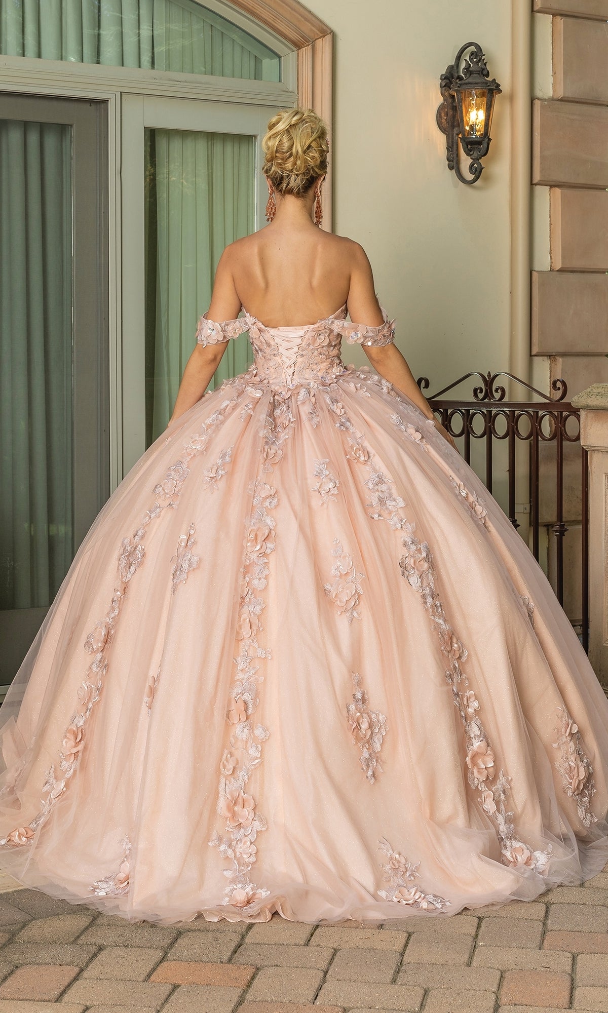 A woman stands outdoors with her back to the camera, wearing Quinceanera Dress 1687 By Dancing Queen—an off-the-shoulder blush pink gown with floral appliques and a ball gown skirt.