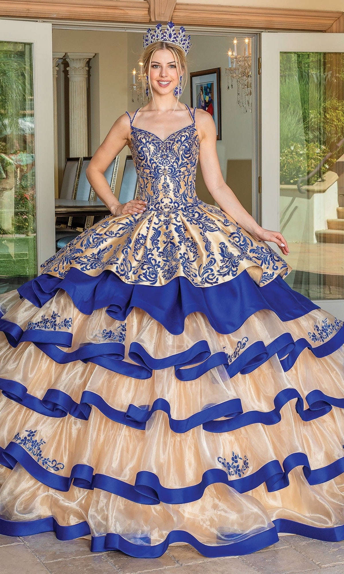 A young woman stands indoors in the Quinceanera Dress 1689 By Dancing Queen in Gold/Royal Blue, featuring intricate embroidery, ruffles, and a tiered ballgown skirt. She wears a matching crown as doors open to a patio—a true Dancing Queen moment.