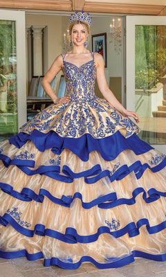 A young woman stands indoors in the Quinceanera Dress 1689 By Dancing Queen in Gold/Royal Blue, featuring intricate embroidery, ruffles, and a tiered ballgown skirt. She wears a matching crown as doors open to a patio—a true Dancing Queen moment.