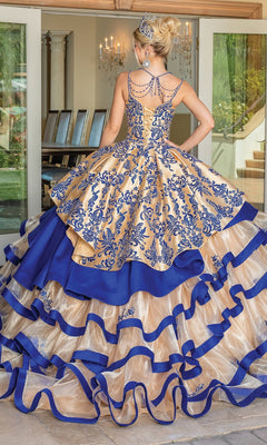 Dressed in the Quinceanera Dress 1689 By Dancing Queen—a gold and blue gown featuring intricate embroidery, a tiered ballgown skirt, and a sparkling tiara—this individual stands with their back to the camera before glass doors, embodying graceful elegance.