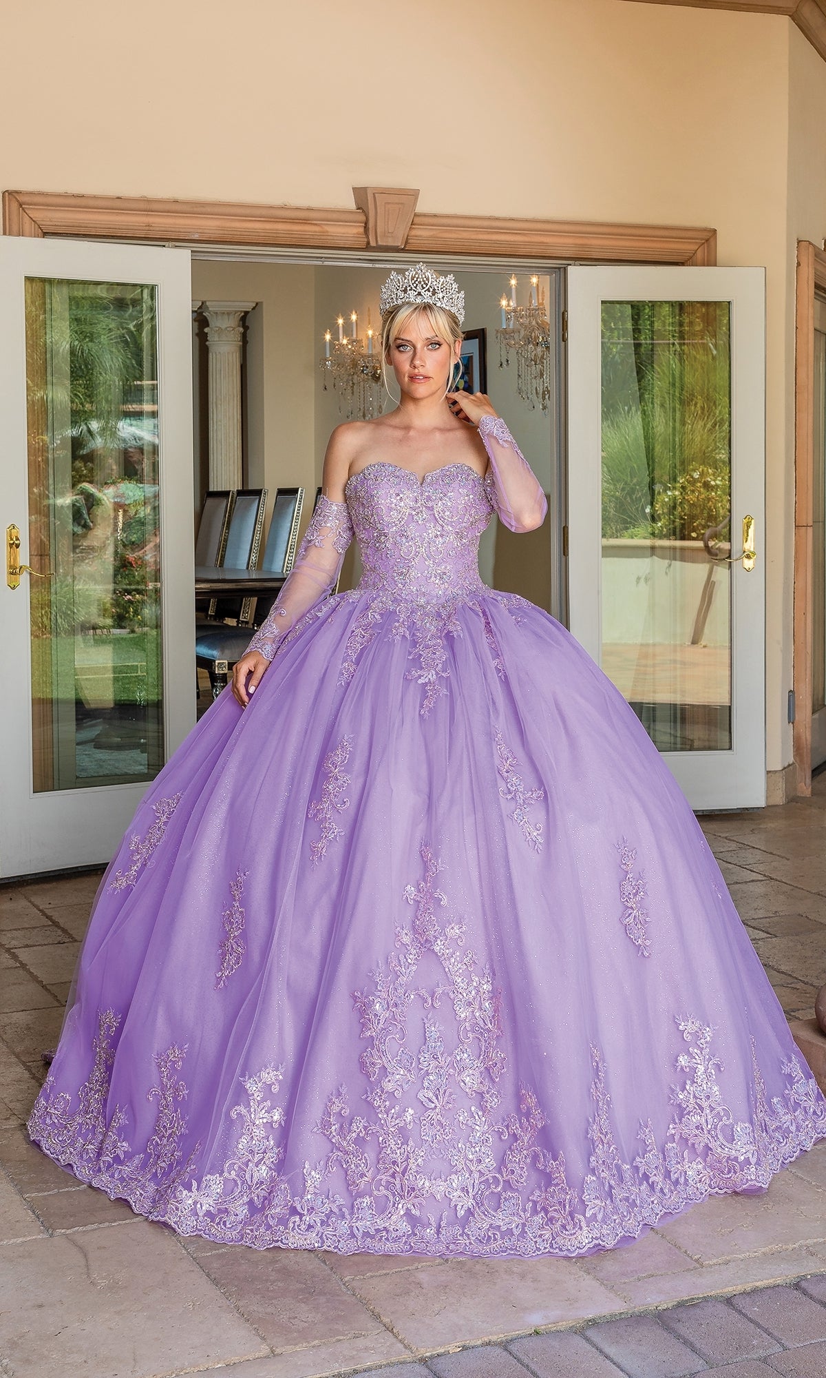 A woman wearing the Quinceanera Dress 1691 by Dancing Queen, a lavender strapless ball gown with intricate embroidery, stands in front of open glass doors and wears a tiara.
